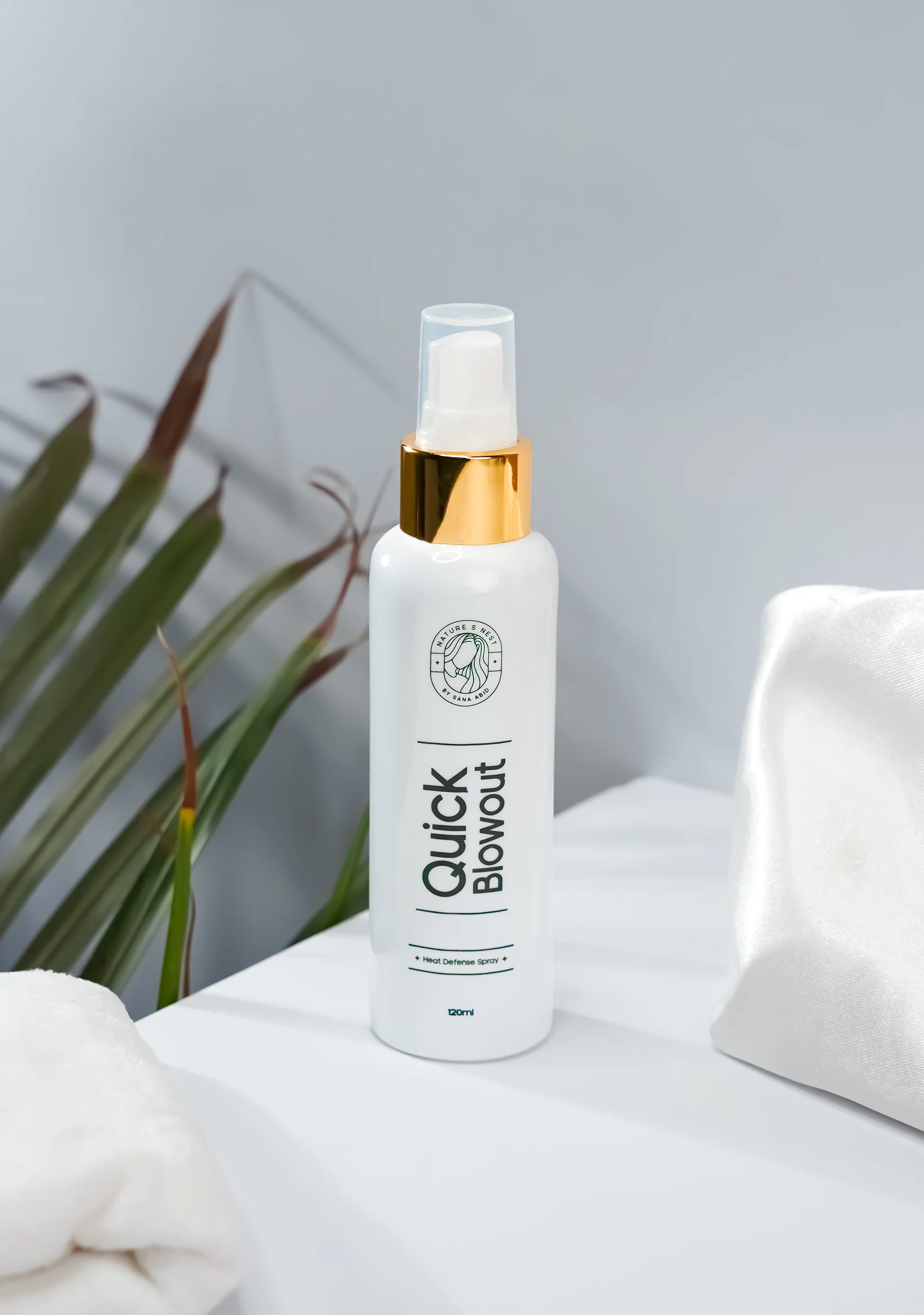 Quick Blowout Heat Defence Spray bottle on a white surface with palm leaves in the background.